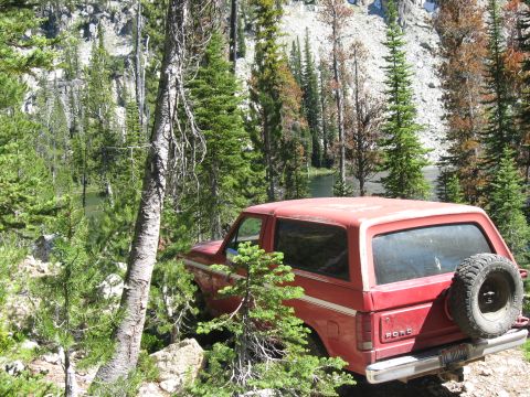 1981 Ford Bronco - A red 1981 Ford Bronco is parked on a rocky trail surrounded by pine trees and a lake in the background.