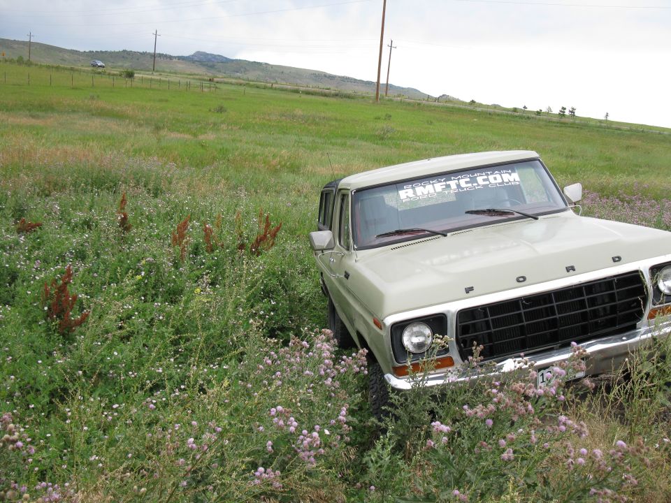 1978 Ford Bronco parked in tall grass with a mountain in the background. "RMFTC.COM" sticker on windshield