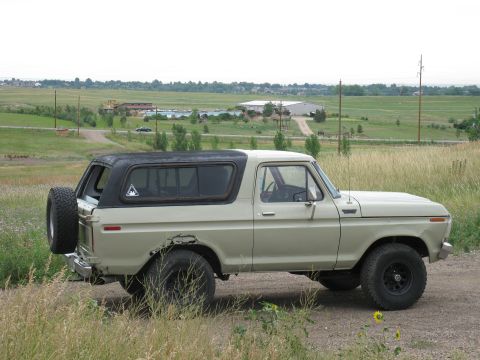 1978 Ford Bronco - A tan 1978 Ford Bronco with a black hardtop is seen from the side, parked on a dirt road with a rural landscape in the background.