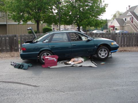 1992 Ford Taurus - A person works on the underside of a green 1992 Ford Taurus sedan, which is parked with its trunk open.