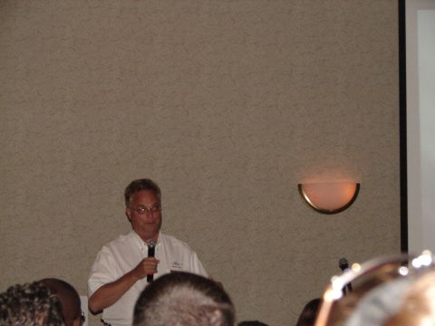 1992 Ford Taurus - A man addresses an audience at a convention, with a wall and a light fixture visible in the background.