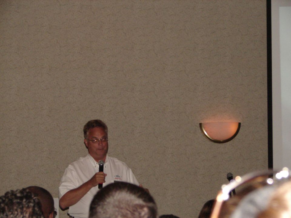Man in white shirt speaking into a microphone at a convention, with a wall and sconce behind him