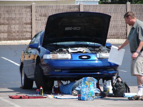 1992 Ford Taurus - A mechanic works on the underside of a blue 1992 Ford Taurus SHO, while another person stands nearby holding a white tray.