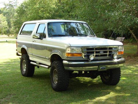 1995 Ford Bronco - This lifted 1995 Ford Bronco features a two-tone paint scheme and off-road tires, parked on a grassy lawn with trees in the background.