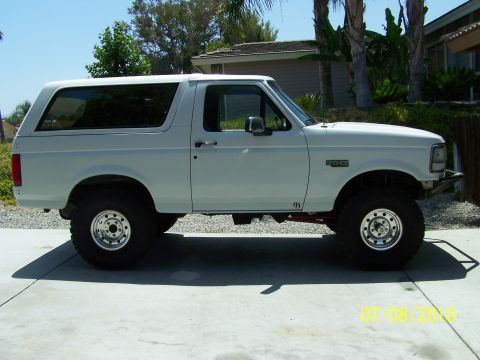 1995 Ford Bronco - A white Ford Bronco is shown from the side, parked on a concrete driveway with a house and palm trees in the background.