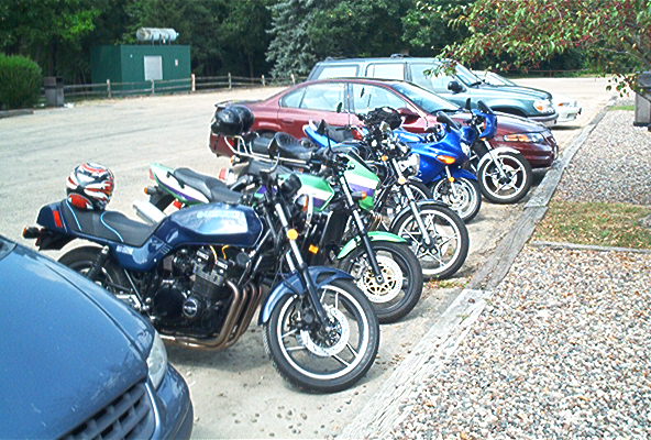 Row of parked motorcycles and sportbikes lined up along a paved lot with cars behind