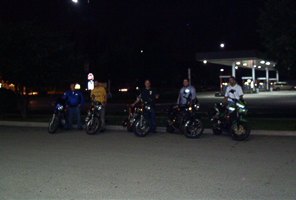 Five people with motorcycles in a dark parking lot at night near a gas station