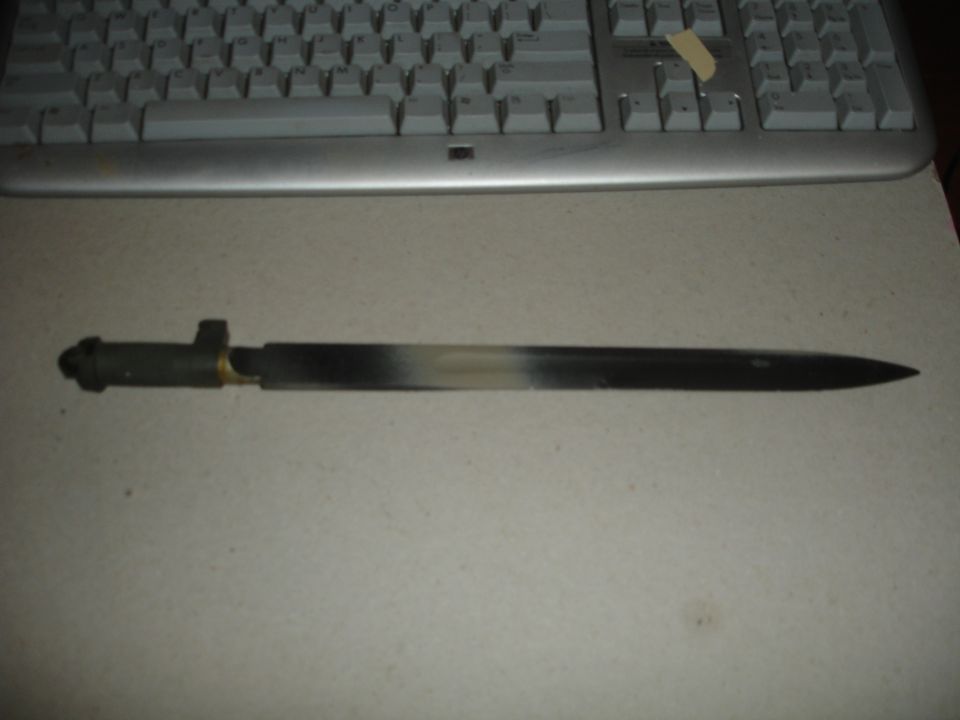 Bayonet with a patterned blade and a dark handle, resting on a surface in front of a keyboard