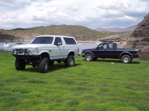1993 Ford Bronco - Two modified trucks are parked on a grassy hill overlooking a lake with boats in the distance.