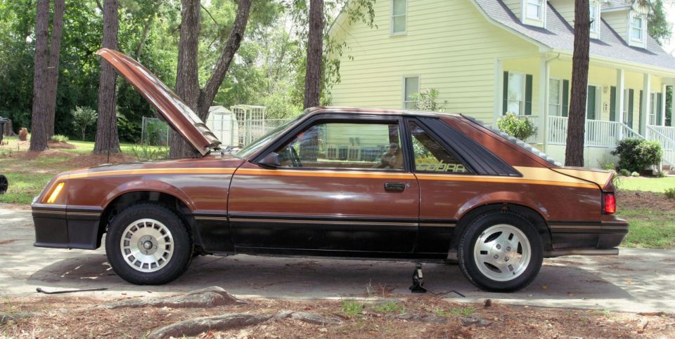 Brown 1980 Ford Mustang Cobra coupe with hood open, parked outside a house