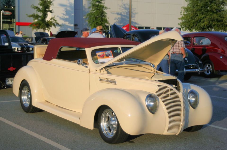 Cream convertible with maroon top, hood open, parked at car show. Other classic cars visible