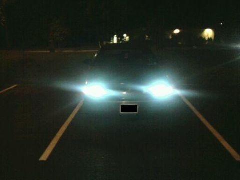 1992 Ford Crown Victoria - A car's bright headlights illuminate the darkness of a parking lot at night.