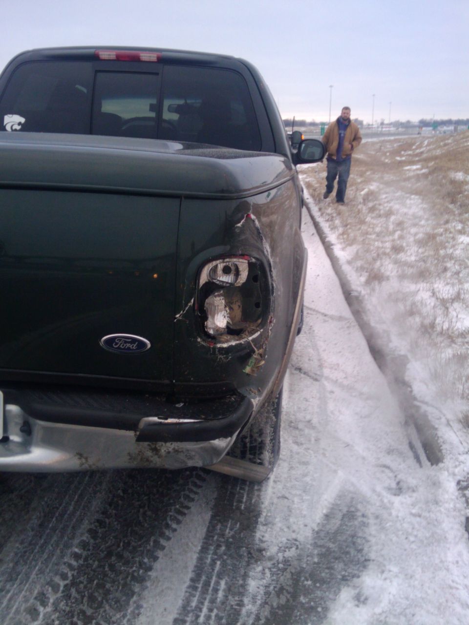 Dark green Ford pickup truck rear view with damaged taillight and a man walking in the background