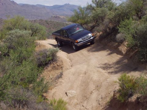 1993 Ford Bronco - A dark blue pickup truck navigates a steep, dusty trail in a mountainous desert environment.