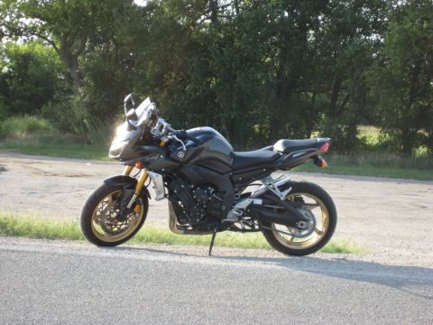 2008 Yamaha FZR1000 - A dark motorcycle is parked on the shoulder of a road, with a wooded area behind it.