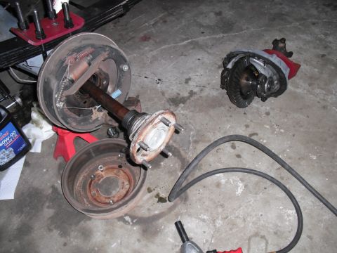 1991 Ford Bronco - Disassembled vehicle axle and differential components are shown on a garage floor, with a bottle of gear oil and tools present.