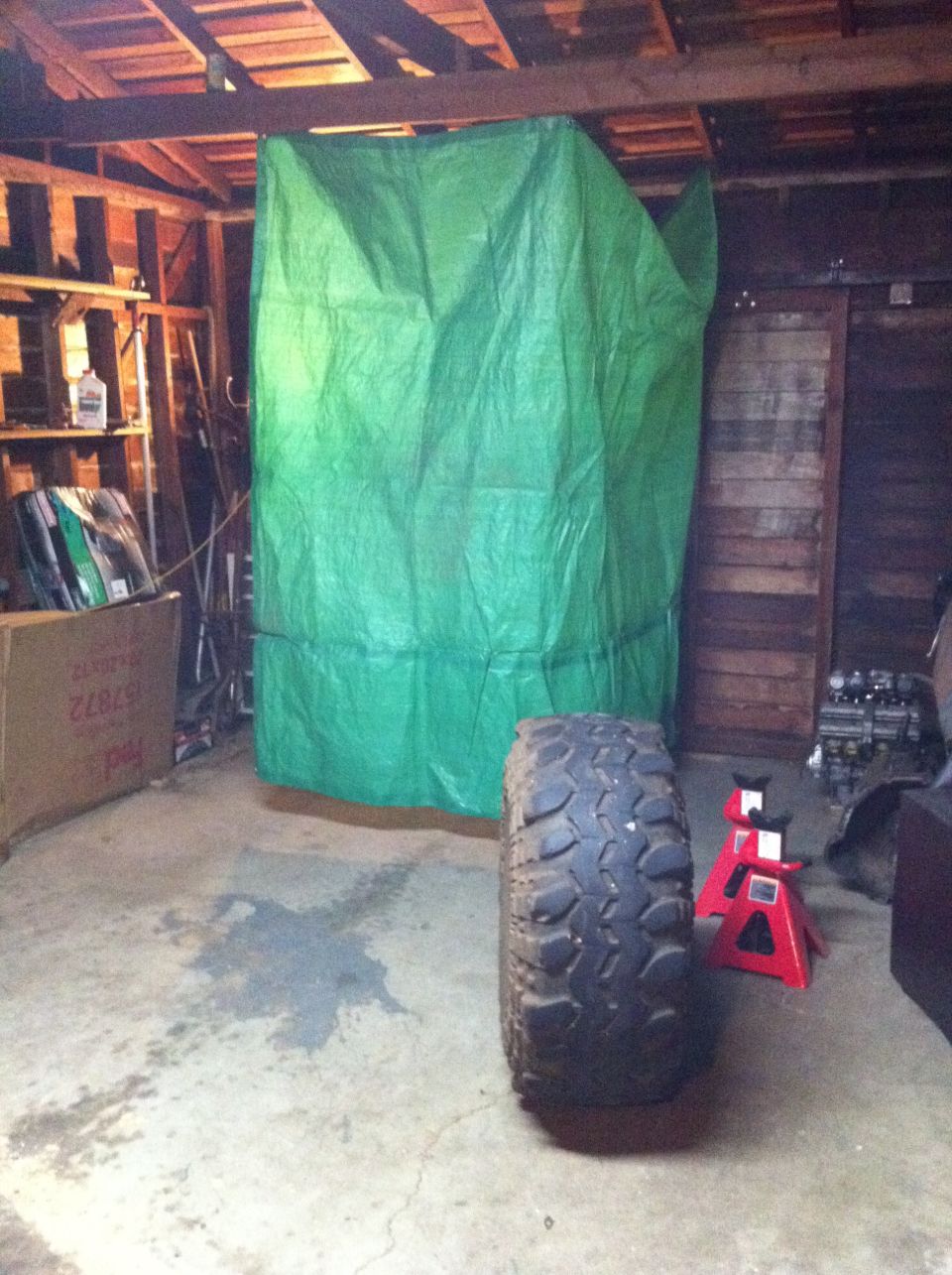 Large off-road tire in foreground, green tarp covering an object in a garage. Red jack stands visible