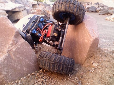 1996 Ford Bronco - A remote control rock crawler is stuck on its side between large rocks on a dirt and gravel terrain.