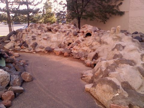 1996 Ford Bronco - A rock formation with multiple openings is situated next to a paved path, with a car visible in the distance.