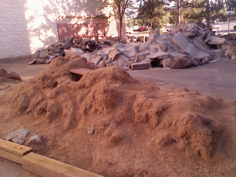 Dirt mounds and rocks in an outdoor area, with a building and trees in the background. A watermark reads SUPERMOTORS