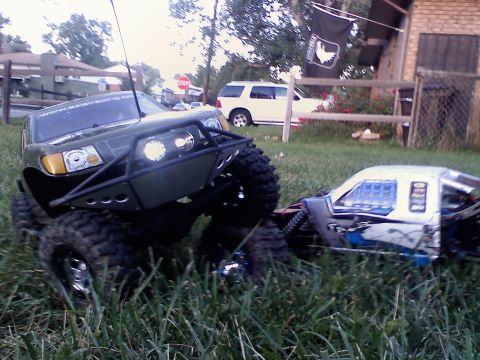 1996 Ford Bronco - Two remote control rock crawlers are parked on a grassy area with a house and fence in the background.