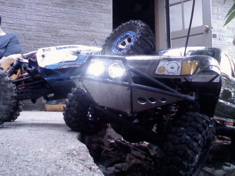 1996 Ford Bronco - Two RC rock crawlers are positioned on a cracked concrete surface, with one in the foreground a prominent bumper and lights.