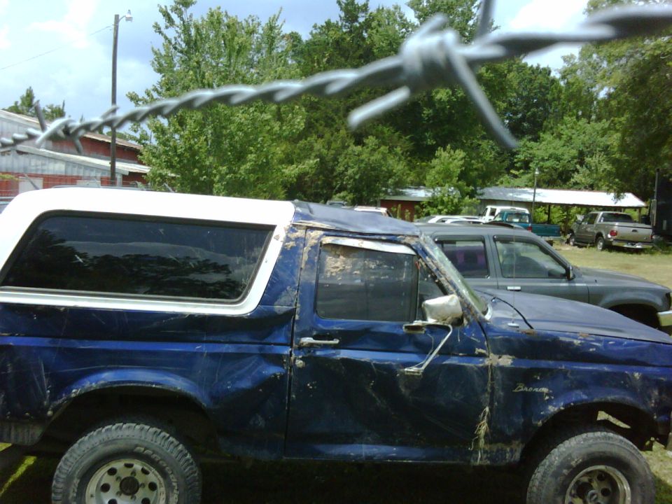 Damaged blue Ford Bronco with white hardtop, side view behind barbed wire