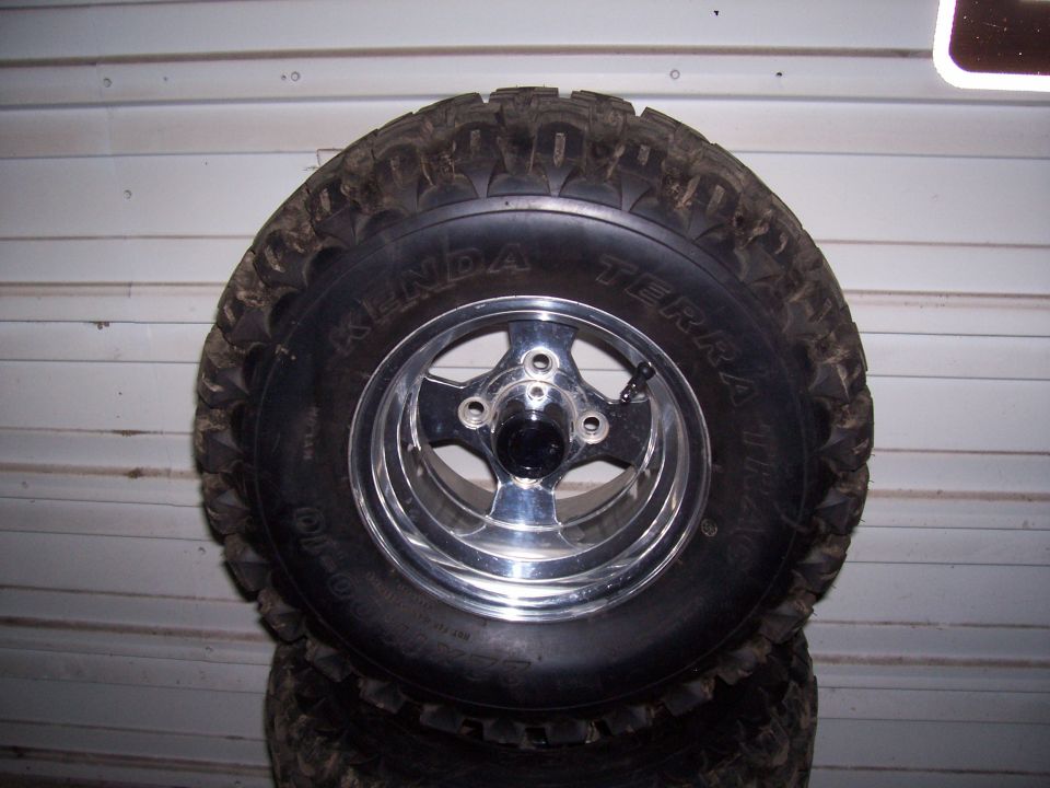 Tire with Kenda Terra Trac branding on a chrome five-spoke wheel, stacked on other tires
