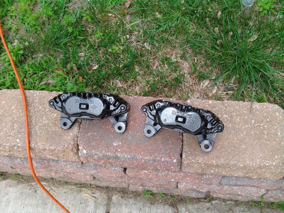 Two gloss black Mazda brake calipers freshly painted, sitting on a brick wall next to grass