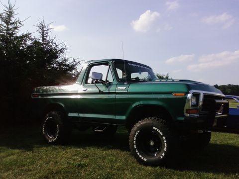 1979 Ford Bronco - Lifted green Ford Bronco with aftermarket wheels and tires sits on a grassy field under a partly cloudy sky.