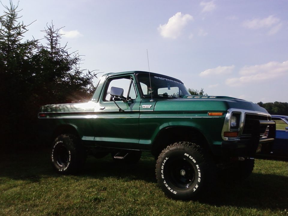 Green lifted Ford Bronco parked on grass, trees in background. "SUPERMOTORS" text on windshield banner