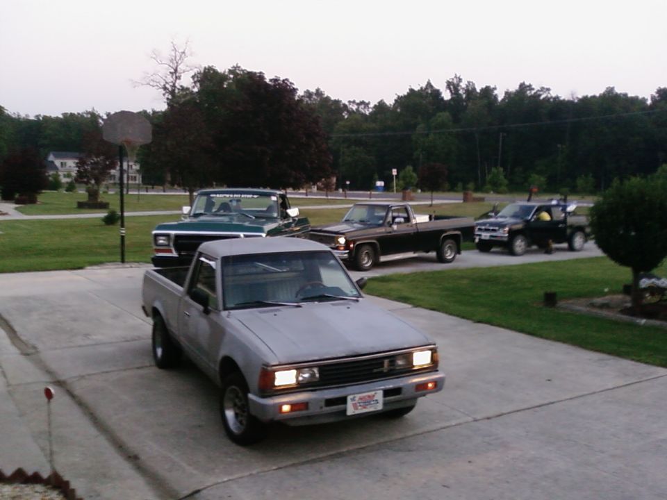 Four pickup trucks parked in a driveway, front one is gray, behind it is a green truck, then a brown truck, and a black truck in the back