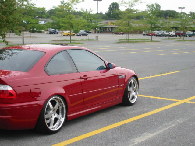 Red coupe with aftermarket wheels parked in a lot. The car has a side vent and appears to be a BMW M3