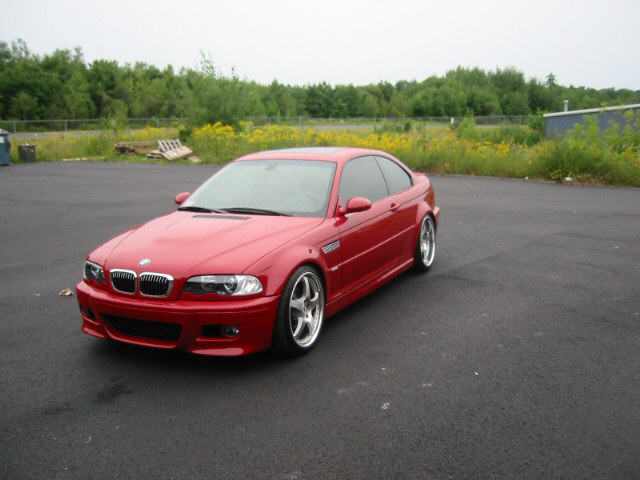 Red BMW E46 M3 coupe parked on asphalt with trees and yellow flowers in background