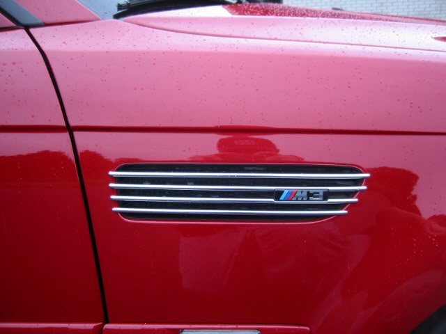 Close-up of a red car's fender showing a horizontal vent with an M3 badge