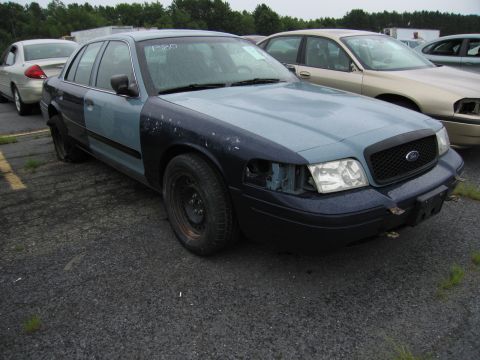 2001 Ford Crown Victoria - A damaged 2001 Ford Crown Victoria with mismatched fenders sits in a lot with other vehicles.