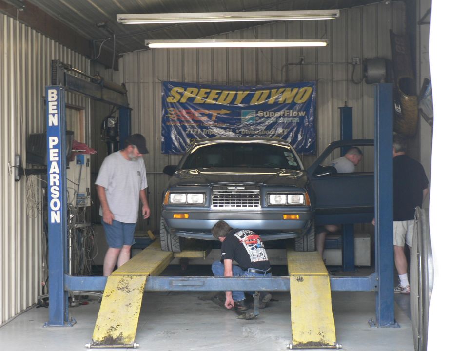 Grey 1980s Ford Thunderbird strapped on a blue four-post dyno under a Speedy Dyno banner