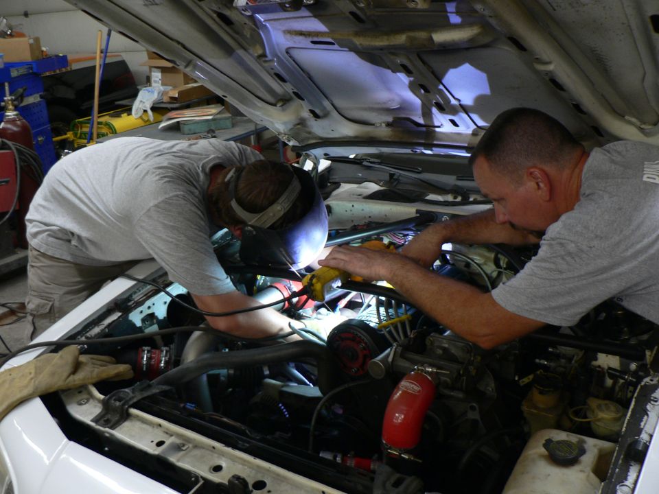 Two men welding in the engine bay of a white car with red silicone intake hose