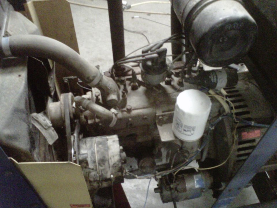 Close-up of an engine with a generator attached. Shows hoses, wires, and an oil filter