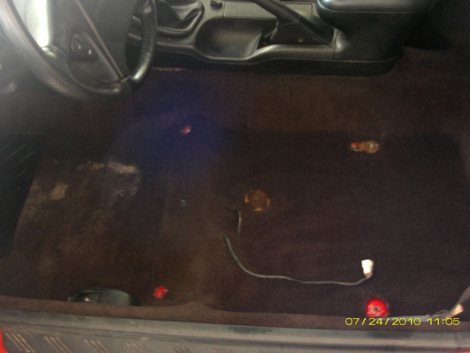 Car interior showing floor carpet, steering wheel, and center console. Some red and yellow lights are visible on the floor
