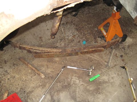 1979 Ford Bronco - Close-up of a rusty leaf spring and shock absorber, surrounded by various tools and an orange jack stand.