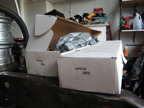 1986 Ford Bronco - Two boxes containing parts labeled EXP4702 are placed on a vehicle's frame in a garage setting.