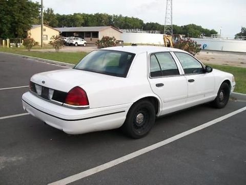 1999 Ford Crown Victoria - A white sedan is parked in a lot with buildings and trees in the background.