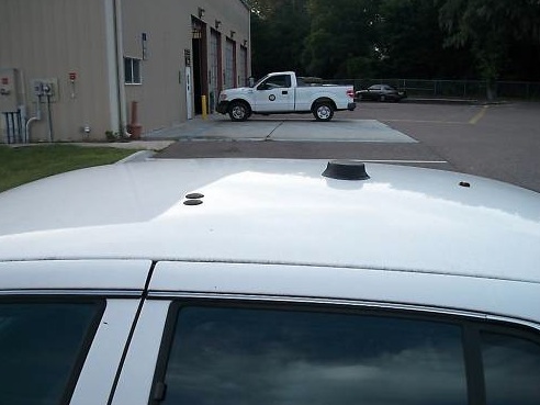 Top view of a white sedan's roof with an antenna; a white pickup truck is parked in front of a building in the background