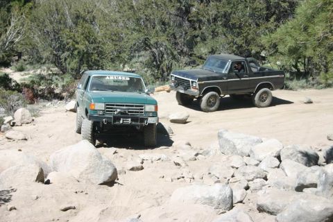 1991 Ford Bronco - Two SUVs are parked on a dirt road, surrounded by trees and rocky terrain.