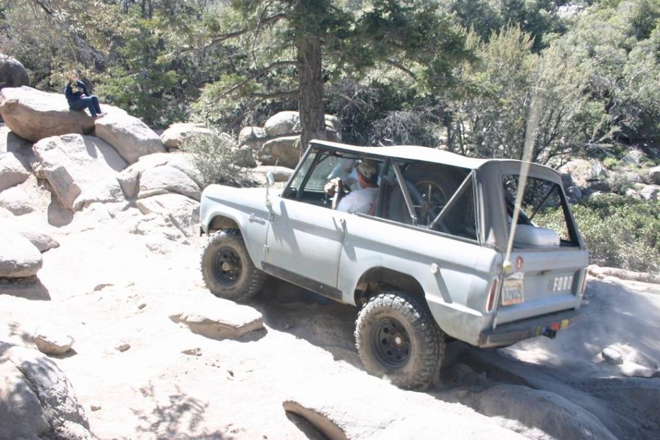 Gray Ford Bronco driving on a rocky trail. A person sits on a rock nearby