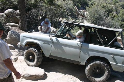 1991 Ford Bronco - A gray Ford Bronco navigates a challenging rocky terrain with assistance from onlookers.