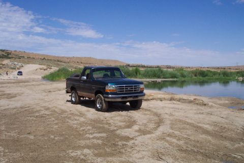 1995 Ford Bronco - A two-door Ford F-150 pickup truck is parked on a dirt path next to a calm body of water under a blue sky.