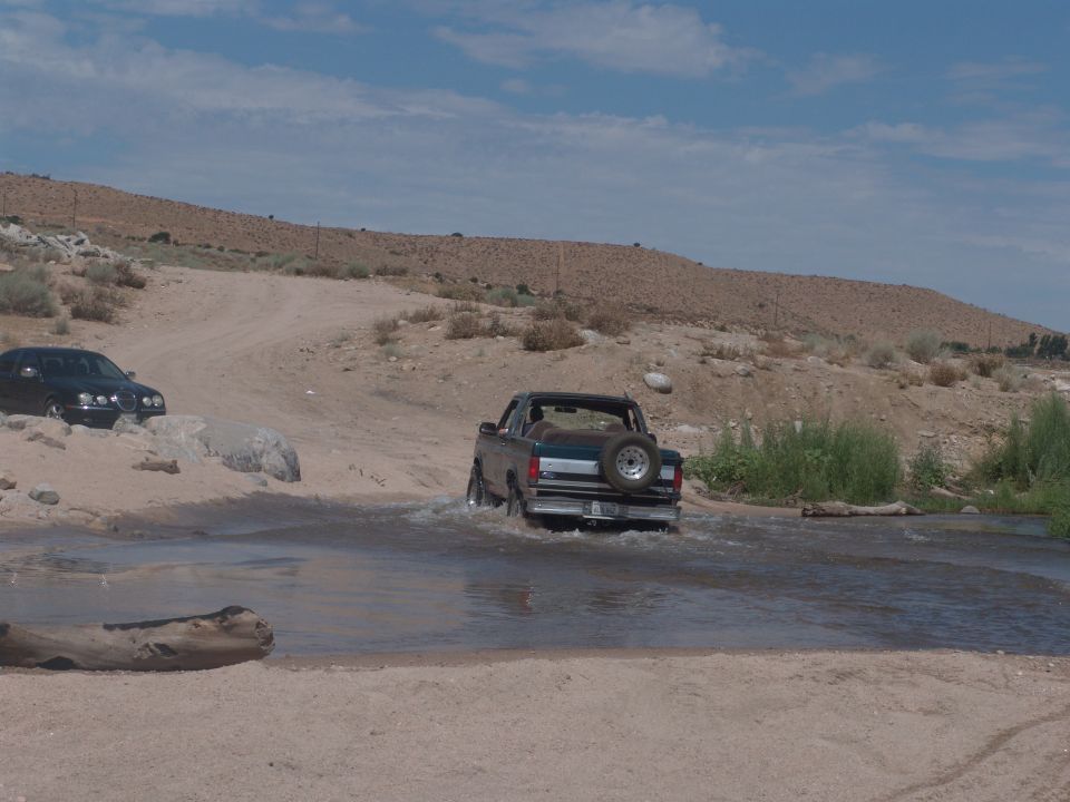 Rear view of a 1995 Ford Bronco driving through water, with a black sedan on the left