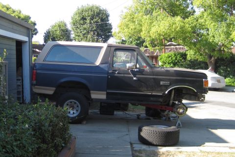1989 Ford Bronco - A 1989 Ford Bronco is parked in a driveway with its front end elevated for maintenance, showing the suspension and missing wheels.